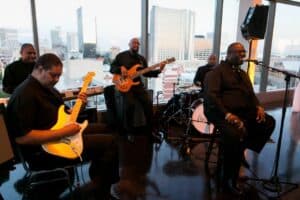 A five-piece band performs indoors with city buildings visible through large windows, as seen in the original post. One man sings into a microphone while others play electric guitar, bass, and drums; all are dressed in black.