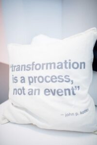 A white pillow featuring grey text that reads, “transformation is a process, not an event — john p. kotter,” as shown in the original post.