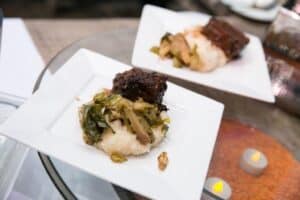 Two square white plates, each with braised meat, mashed potatoes, and sautéed greens, are arranged on a glass table beside small decorative LED candles in this WM Events dining setup.