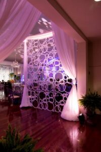 A modern event space designed by WM Events, featuring sheer white drapes, a decorative circular divider, glossy wooden floor, and purple ambient lighting. Lush ferns and elegant dining tables are visible in the background.